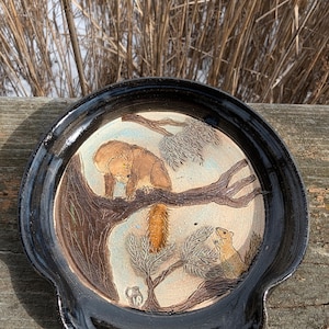 May include: A ceramic dish with a black rim and a white interior. The dish features a hand-painted scene of a red fox on a tree branch with two squirrels below. The dish is perfect for serving snacks or holding small items.
