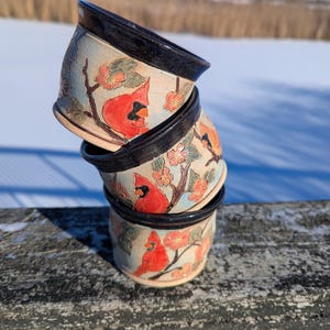 May include: Stacked ceramic pots with a light blue background, black rims, and a hand-painted design. The design features red cardinals, orange flowers, and green leaves on branches. The pots are stacked on a weathered wooden surface.