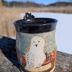 May include: Handmade ceramic mug with a black rim and handle. The mug features a detailed relief of a white owl with yellow eyes, set against a backdrop of a brick wall and a red and gold design. The mug is set on a wooden surface.