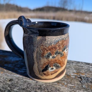 May include: A handcrafted ceramic mug with a dark brown handle and rim. The mug features a detailed relief carving of raccoons in shades of brown and tan. The mug is set against a natural outdoor backdrop.