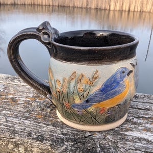 May include: A ceramic mug with a dark brown rim and handle, featuring a hand-painted bluebird design. The mug has a light blue background with tall grass and flowers. The base is a light beige color.