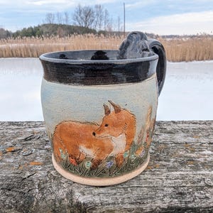 May include: A handmade ceramic mug with a fox design. The mug features a brown fox in a field of flowers, with a black rim and handle. The mug is sitting on a weathered wooden surface.