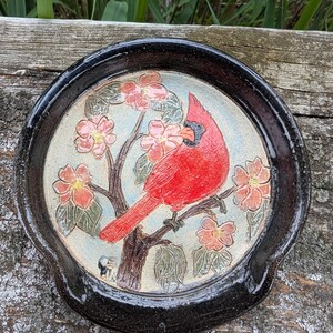 May include: A round ceramic spoon rest featuring a glossy black rim and a central illustration of a bright red cardinal bird perched on a brown tree branch surrounded by light pink flowers with yellow centers against a pale blue background.
