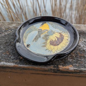 May include: A round, black ceramic spoon rest with a hand-painted design. The design features a yellow and black bird perched on a sunflower. The background is light blue.
