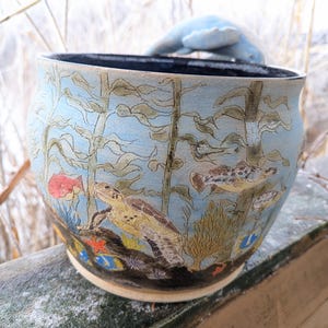 May include: A ceramic bowl with an underwater scene. The bowl features a light blue background with illustrations of seaweed, fish, and a sea turtle. A light blue dolphin-shaped handle is attached to the rim.
