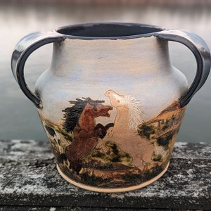 Puede incluir: Un jarrón de cerámica con dos asas, que presenta una escena pintada a mano de dos caballos en un campo. El jarrón tiene un esmalte marrón claro con un lavado azul y blanco.