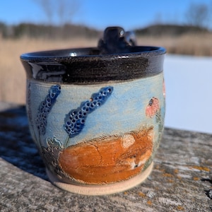May include: Handmade ceramic mug with a fox design. The mug features a blue sky background with floral and plant details. The fox is orange and brown. The rim and handle are black. The mug sits on a light beige base.