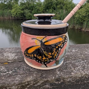 May include: A ceramic honey pot with a glossy black lid and a matching black rim. The body features a textured, hand-painted orange and black butterfly design against a soft pink background. A light-colored wooden honey dipper rests inside the pot.