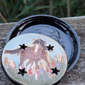 May include: A ceramic horse-themed decorative piece with a lid. The lid features a brown horse illustration with black star cutouts. The base is black and glossy. The background shows blurred greenery.