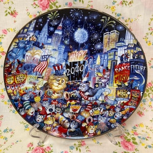 Peut inclure: Une assiette décorative représentant une célébration du Nouvel An avec des chats de dessins animés dans un paysage urbain. L'assiette a un fond bleu foncé avec des feux d'artifice, des bâtiments et le texte "Happy 2000". L'assiette est soutenue par un support en plastique transparent.