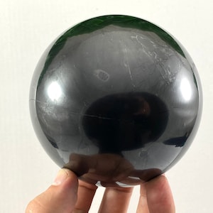 Può includere: Una sfera di ossidiana nera lucidata, un tipo di cristallo, è tenuta nella mano di una persona. La sfera è liscia e riflettente, con un leggero bagliore.