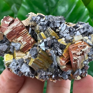 May include: A mineral specimen featuring a cluster of metallic cubic crystals in shades of copper, gold, and silver. The structure displays sharp, geometric facets with a reflective, high-luster finish, interspersed with dark gray, irregular mineral deposits.