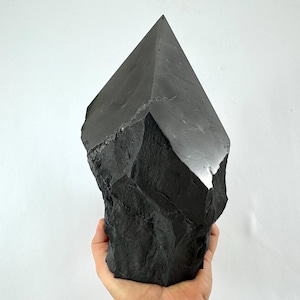 May include: A large, dark gray obsidian crystal point with a rough, textured base and a smooth, polished top. The crystal is held in a hand against a white background. This natural stone is a decorative item.