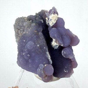 Puede incluir: Primer plano de un espécimen mineral de ágata de uva. La pieza presenta un color púrpura intenso con formaciones redondeadas en forma de burbujas. La superficie tiene un aspecto texturizado, con algunas zonas más claras, casi blancas.