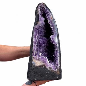 May include: A large, tall amethyst geode featuring a deep purple, crystalline interior cavity set within a dark, rough, natural rock exterior. The structure is elongated and pointed at the top, showcasing a dense cluster of jagged, translucent purple quartz crystals that contrast sharply against the solid, dark, matte stone base.