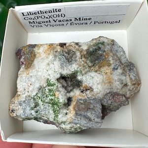 May include: A rough-hewn mineral specimen of Libethenite, Cu2(PO4)(OH), from Miguel Vacas Mine, Portugal, displayed in a white box. The stone features a mix of green, white, gray, and brown hues. The text "Libethenite" is visible.