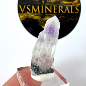 Può includere: Un cristallo di quarzo trasparente con una punta viola, esposto su un supporto in acrilico trasparente. Il cristallo ha una consistenza ruvida e una base scura. Lo sfondo include un logo circolare nero e oro con il testo "VSMINERALS".