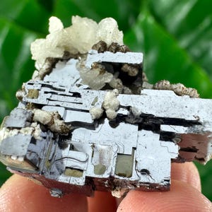 May include: A close-up of a metallic gray mineral specimen with a layered, geometric structure. The surface has a reflective quality, with patches of lighter, crystalline formations and brown inclusions. The background is blurred green.