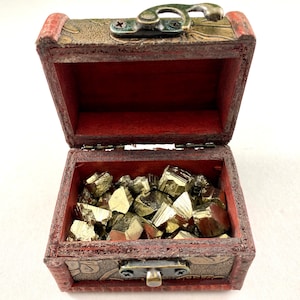 Puede incluir: Un pequeño cofre del tesoro de madera abierto, lleno de cristales de pirita dorada. El cofre es marrón con interior rojo y detalles metálicos decorativos. Los cristales tienen brillo metálico.