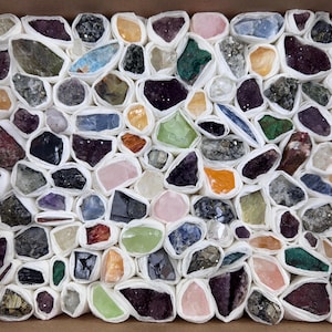 Pode incluir: Uma coleção de vários espécimes minerais brutos e cristais, cada um embrulhado individualmente em papel branco, dispostos em uma grade densa dentro de uma caixa quadrada. As pedras exibem uma ampla gama de cores, incluindo roxo, verde, laranja e azul.