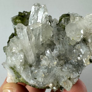 May include: A cluster of clear quartz crystals with green inclusions. The crystals are arranged in a cluster, with some of the crystals pointing upwards. The crystals are surrounded by a matrix of green material.