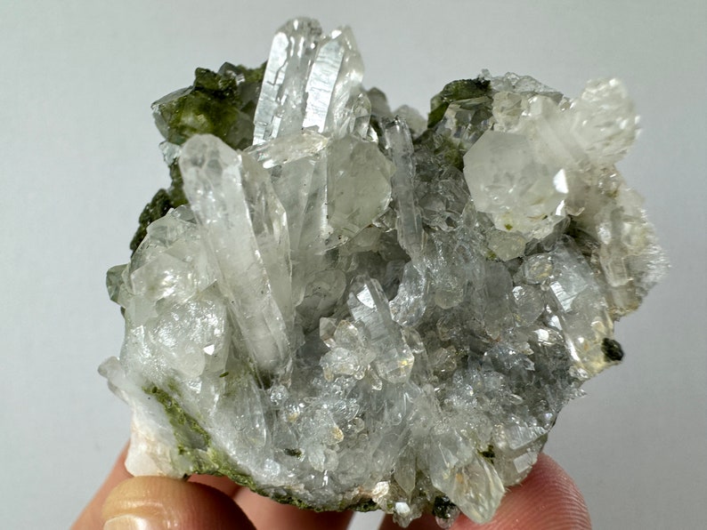 May include: A cluster of clear quartz crystals with green inclusions. The crystals are arranged in a cluster, with some of the crystals pointing upwards. The crystals are surrounded by a matrix of green material.