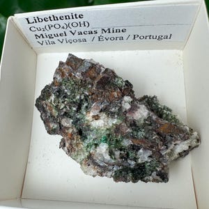 May include: A specimen of Libethenite, a copper phosphate mineral, displayed in a white box. The mineral has a rough, textured surface with shades of brown, green, and white. The box label includes the mineral's name, chemical formula, and origin from Miguel Vacas Mine, Portugal.
