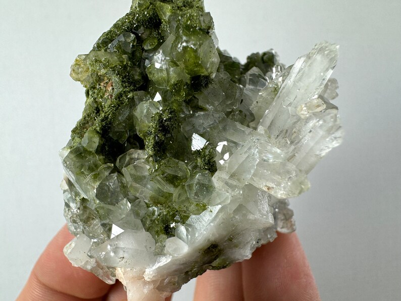 May include: A cluster of clear quartz crystals with green epidote inclusions. The crystals are arranged in a random pattern and have a natural, rough texture.