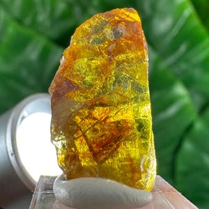 Peut inclure: Une pierre précieuse brute, translucide, de couleur jaune et orange, avec une texture cristalline et irrégulière. La pierre présente des fractures et des inclusions internes, éclairée par une source lumineuse vive sur le côté.