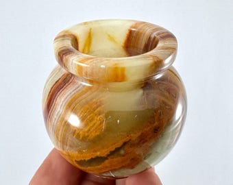 Può includere: Un piccolo vaso rotondo in onice con un'ampia apertura. Il vaso presenta fasce di colori marrone, giallo e crema. La superficie lucida riflette la luce, evidenziando i motivi naturali della pietra. Un oggetto decorativo.