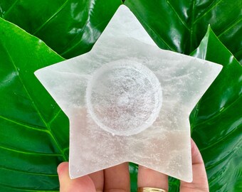 May include: A star-shaped white selenite crystal candle holder with a smooth, translucent surface and a circular indentation in the center. The object features a five-pointed design with a matte, crystalline texture throughout its flat structure.