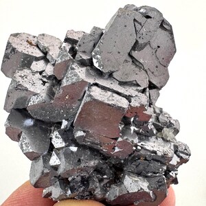 May include: A cluster of metallic gray galena crystals featuring sharp, cubic structures with reflective surfaces. The mineral specimen displays a dense arrangement of interlocking geometric shapes with a dark, lustrous finish and visible crystalline facets throughout the formation.