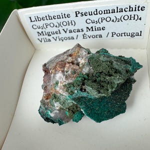 May include: A mineral specimen of Libethenite Pseudomalachite, featuring a mix of green, brown, and gray hues. The mineral is presented in a white box with printed text indicating its composition and origin from Portugal.