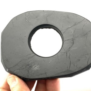 Peut inclure: Une bague en pierre grise avec un trou circulaire au centre. La pierre a une surface rugueuse et texturée et est de forme irrégulière.