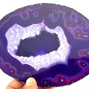 Könnte beinhalten: Eine polierte, oval geformte violette Achat-Scheibe mit konzentrischen Bändern in tiefen Violett- und Magentatönen. Die Mitte enthält einen gezackten, kristallinen Geoden-Hohlraum mit weißen, durchscheinenden Quarzformationen und einem dunklen Kern.