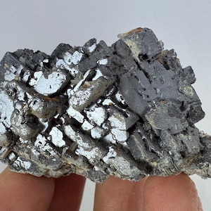 May include: A raw mineral specimen featuring a cluster of metallic, dark gray cubic crystals. The structure displays sharp, geometric edges and reflective surfaces with a dense, heavy appearance, showcasing natural geological formations.