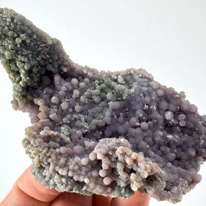 Könnte beinhalten: Ein rohes Exemplar von Traubenachat. Der Stein weist eine Ansammlung kleiner, runder, traubenähnlicher Formationen in den Farbtönen Lavendel, Lila und Grün auf. Die Gesamtstruktur ist uneben und organisch, mit kristallinem Aussehen.