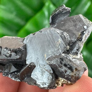 May include: A cluster of dark gray, metallic mineral crystals with sharp, angular facets and a reflective surface. The specimen features multiple interconnected geometric shapes with visible striations and a dense, crystalline structure.