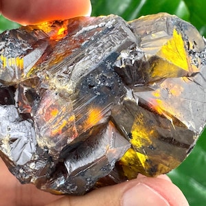 Peut inclure: Un spécimen minéral brut présentant des structures cristallines gris métallique foncé avec des sections translucides de couleur ambre vif. L'amas affiche des facettes angulaires nettes et des formations géométriques complexes et profondes.