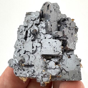 May include: A raw galena mineral specimen featuring a cluster of metallic, silver-gray cubic crystals. The structure displays sharp, geometric edges and a reflective, lead-like surface with small, recessed crevices throughout the formation.