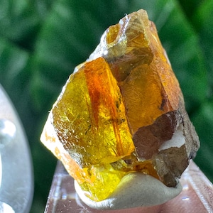 Peut inclure: Un spécimen minéral brut, translucide, de couleur jaune et orange, doté d'une structure cristalline irrégulière. La pierre présente des teintes ambrées profondes, des fractures internes visibles et une texture de surface brute.