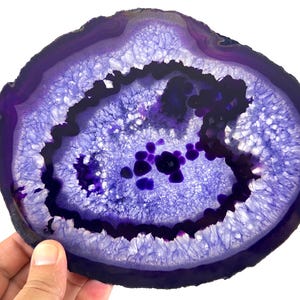 Può includere: Una fetta di agata lucidata con vivaci anelli concentrici viola e un centro cristallino. La pietra presenta tonalità viola intenso sui bordi esterni, che sfumano in una struttura interna cristallina più chiara, testurizzata e scintillante.