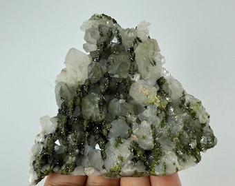 May include: A cluster of clear quartz crystals with green epidote inclusions. The crystals are arranged in a cluster, with some of the crystals pointing upwards. The epidote inclusions are scattered throughout the crystals, giving them a green speckled appearance.