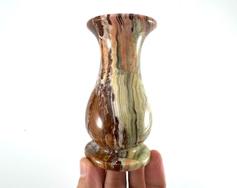 Può includere: Un vaso decorativo con la parte superiore svasata e una base arrotondata. Il vaso è realizzato in pietra con un mix di colori marrone, verde e crema. Il vaso è tenuto in mano.