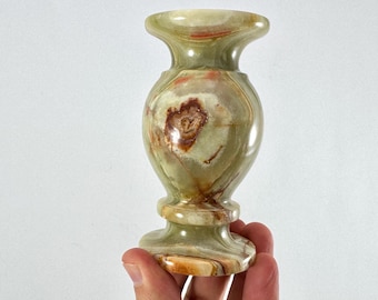 Può includere: Un piccolo vaso decorativo in onice verde e marrone. Il vaso ha una parte superiore svasata, un corpo arrotondato e una base a piedistallo. L'onice presenta motivi naturali e variazioni di colore.