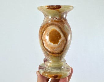 Può includere: Un vaso decorativo in onice con base a piedistallo. Il vaso presenta un bordo svasato e un corpo bulboso con motivi concentrici nei toni del marrone, beige e crema. Il vaso è alto circa 15 cm.