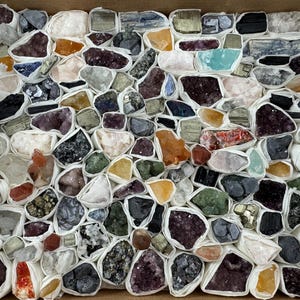 Puede incluir: Una caja llena de una variedad de piedras preciosas y cristales en bruto. Las piedras son de diversos colores, incluyendo púrpura, naranja, verde y transparente, y están envueltas individualmente en papel blanco. La disposición sugiere una colección de minerales.