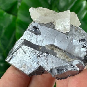 May include: A raw mineral specimen featuring a metallic, dark gray galena base with sharp, cubic crystalline structures. Atop the dark, reflective surface sits a cluster of translucent, off-white calcite crystals with distinct, angular facets.