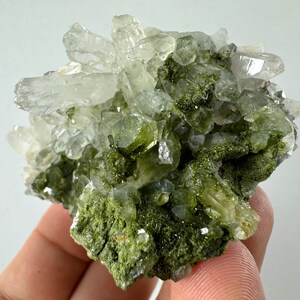 May include: A cluster of clear quartz crystals with green epidote inclusions. The crystals are clustered together in a rough, natural formation.