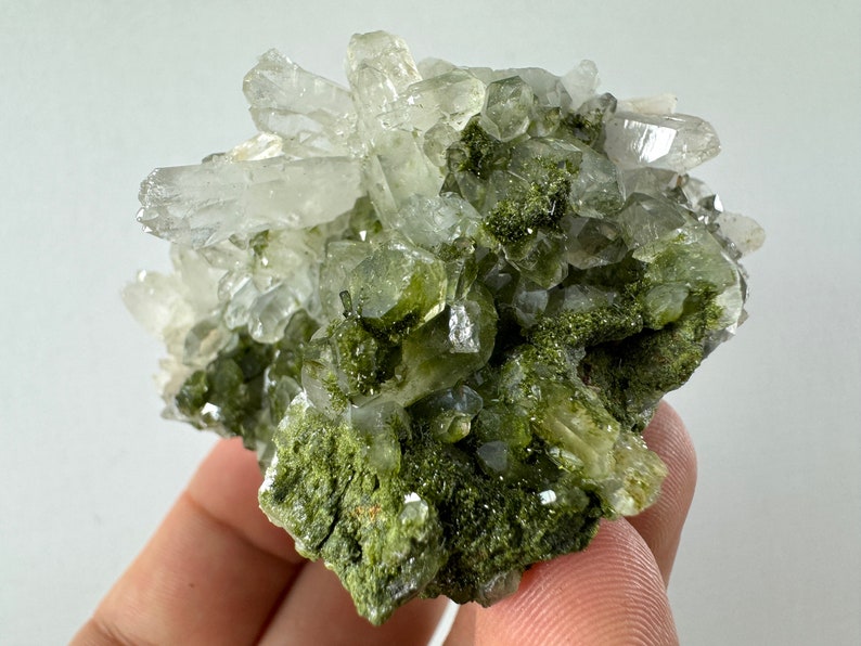 May include: A cluster of clear quartz crystals with green epidote inclusions. The crystals are clustered together in a rough, natural formation.